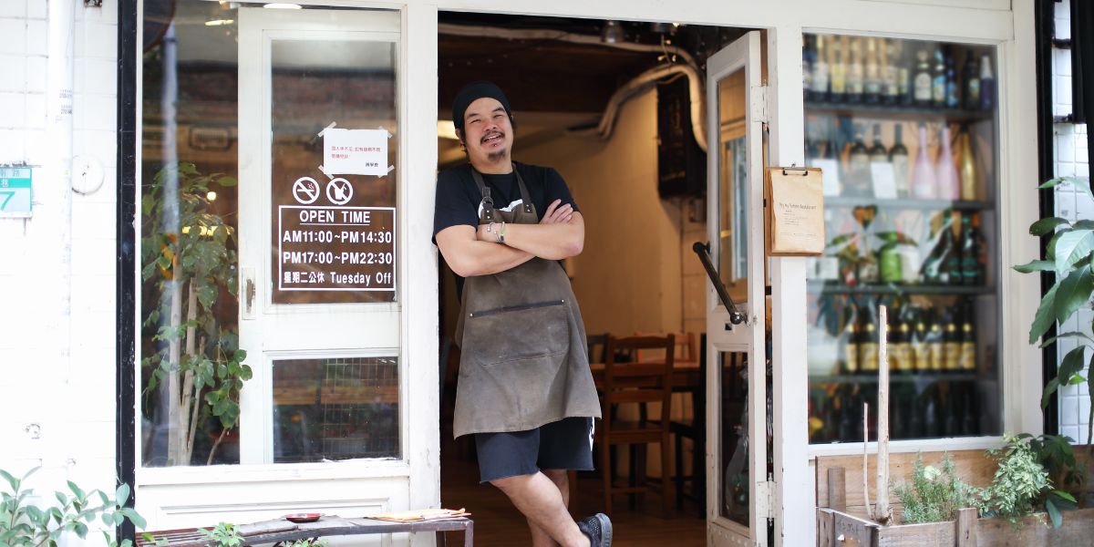 Restaurant owner standing confidently at the entrance of a cozy, modern eatery with arms crossed and a welcoming smile.